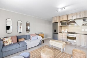 A modern apartment with a grey sofa, a small white bench, and a kitchen with wooden cabinets and stainless steel appliances. - Mayfair 246 (Cape Town)