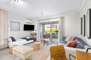 A modern living room with a sofa, ottoman, and a small table. There is a TV mounted on the wall and a balcony with outdoor furniture. - Mayfair 246 (Cape Town)