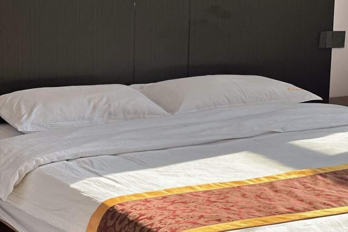 A bed with white linens and a patterned blanket, a dark headboard, and a wall-mounted light switch.