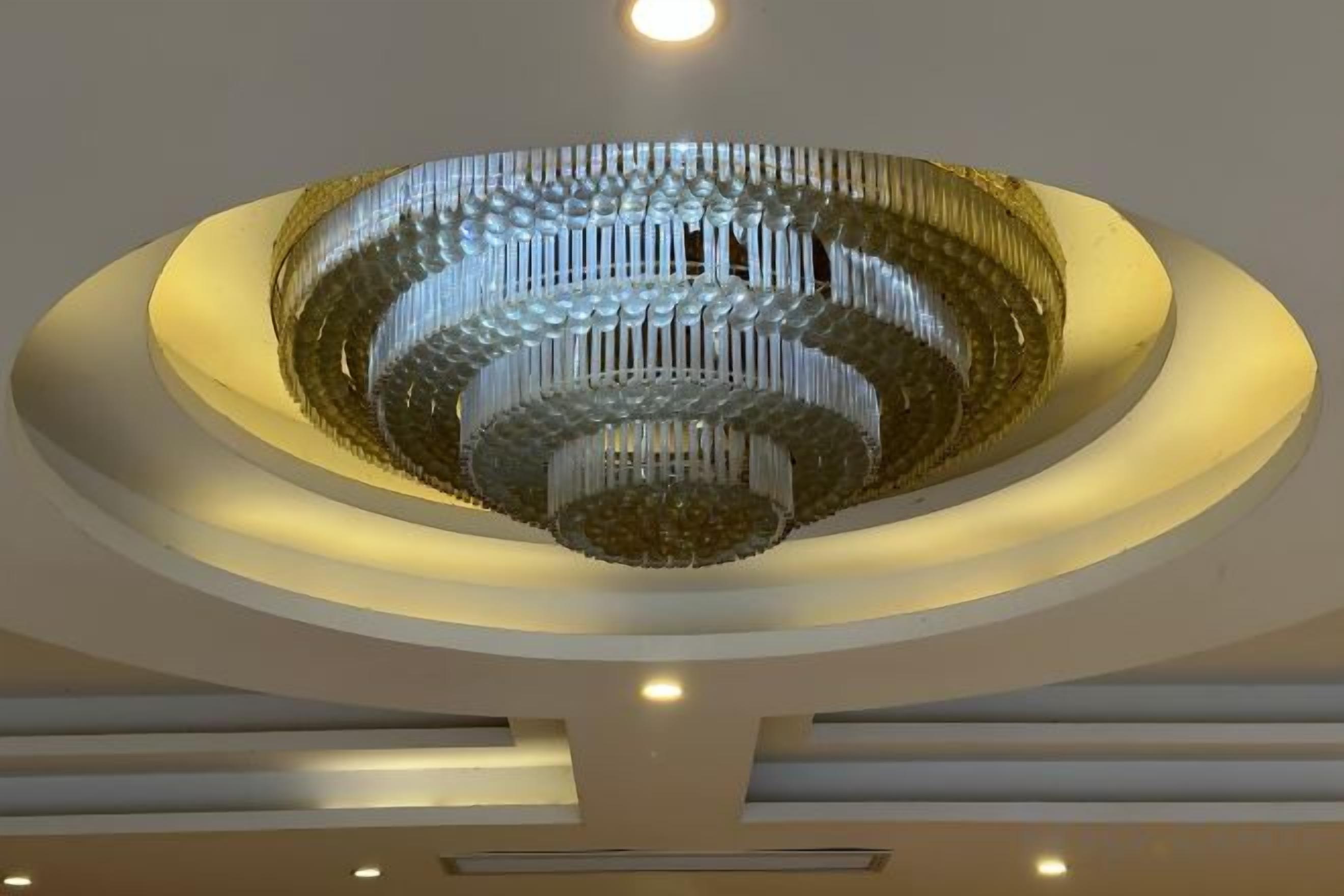 Un grand lustre circulaire, composé de plusieurs niveaux et de nombreuses sources lumineuses, placé au centre d’un plafond doté d’un éclairage encastré.