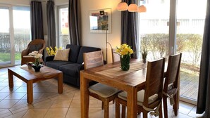 Ein modernes Wohnzimmer mit einer Couch, einem Esstisch und Stühlen, großen Fenstern mit Vorhängen und Blick auf Grünfläche.