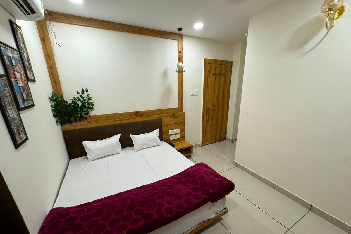 A neatly made bed with white linens and a maroon blanket, a wooden headboard, a bedside table with a lamp, and a potted plant on the wall.