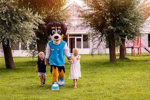 A mascot character in a blue and white jacket walking with two children in a grassy park area.