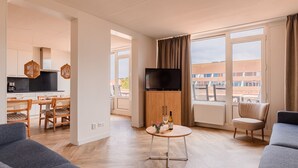 Una sala de estar moderna con televisor, un comedor con mesa y sillas, y ventanas amplias con vista a edificios.