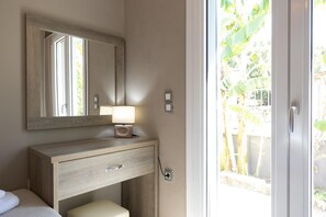 Een badkamer met een houten wastafelmeubel, een spiegel, een bedlamp en uitzicht op een tuin door een glazen deur.