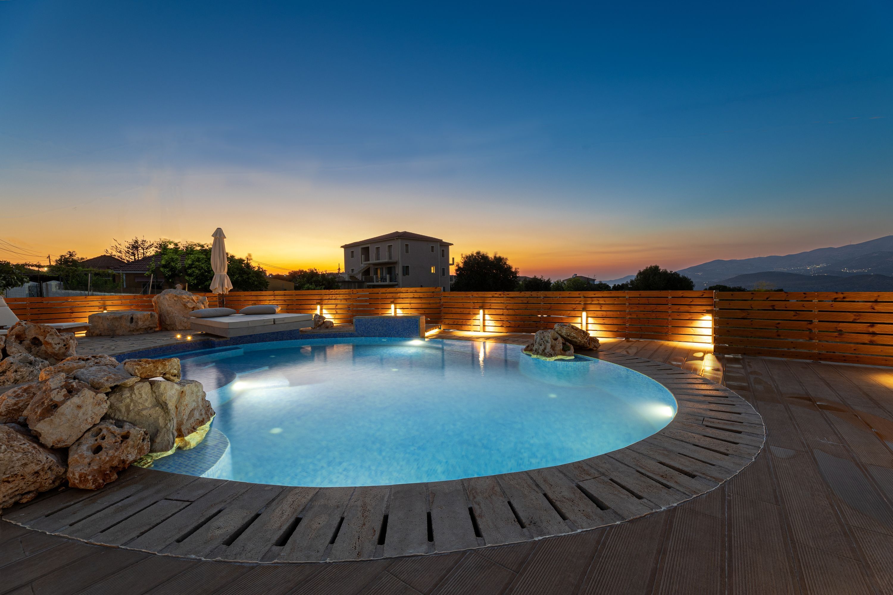 A swimming pool with a wooden deck, surrounded by rocks, and a sunset view in the background.