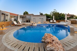 Une piscine entourée de chaises longues et de parasols, elle-même bordée d’une terrasse en bois et d’un bâtiment doté d’un toit en tuiles.