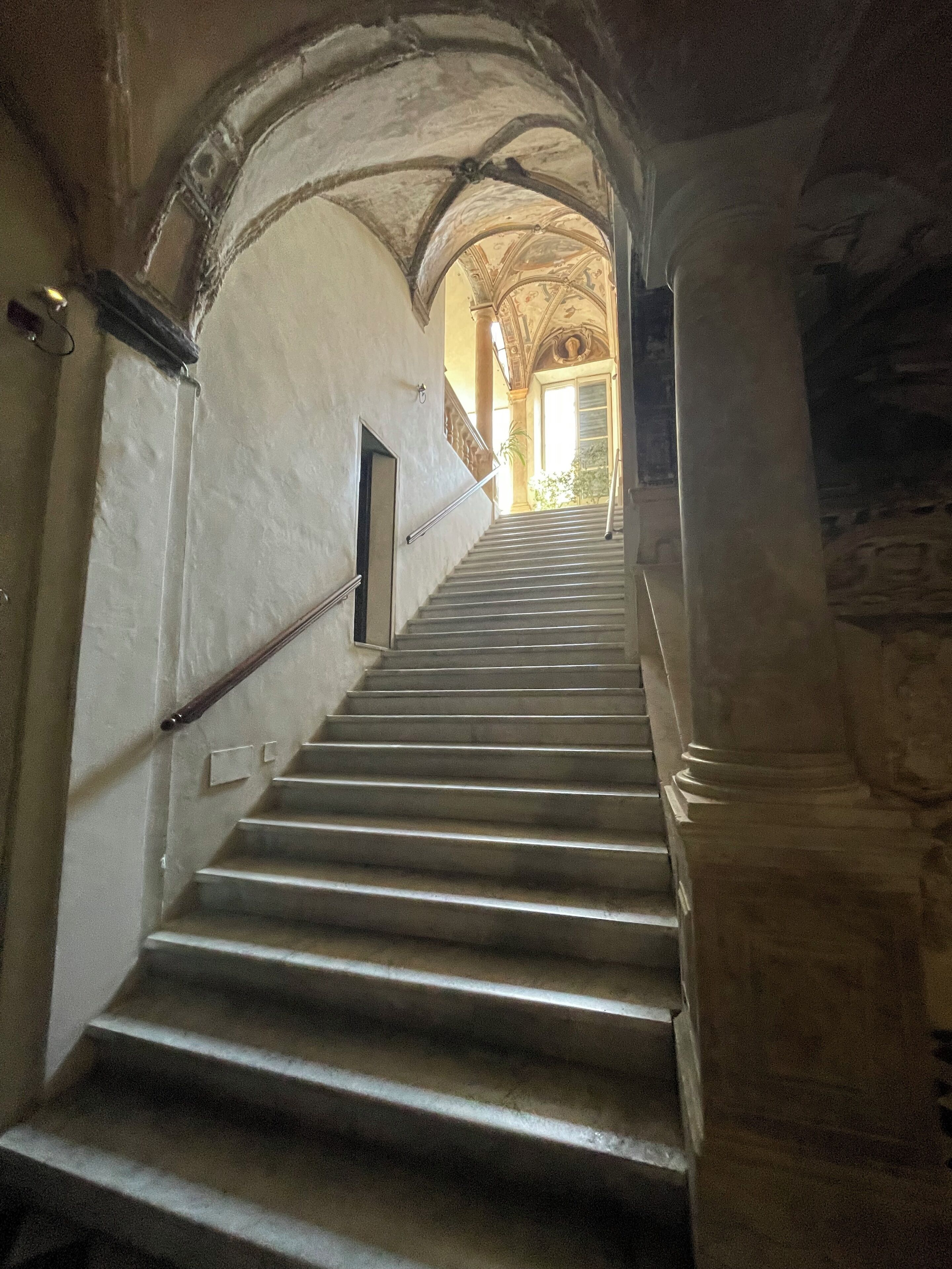A staircase with stone balustrades and arches, leading to a brighter area with a view of greenery.