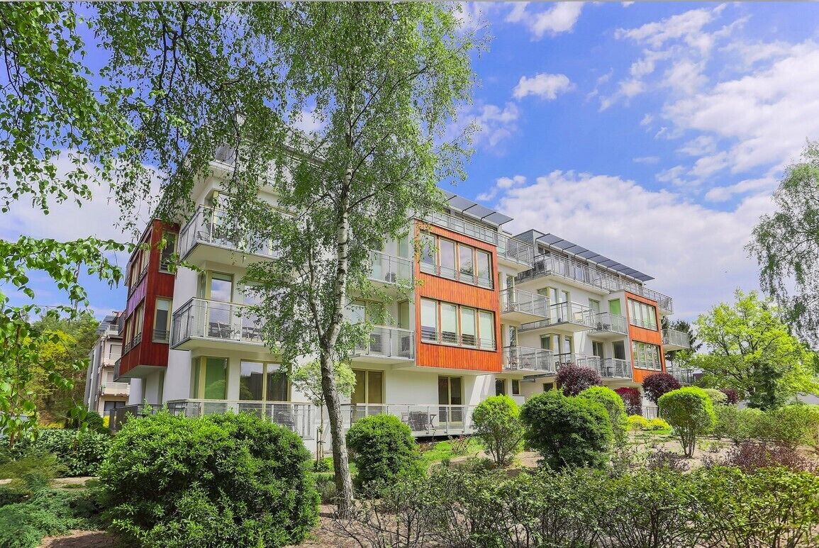 A modern multi-story residential building with balconies, surrounded by lush greenery and trees.