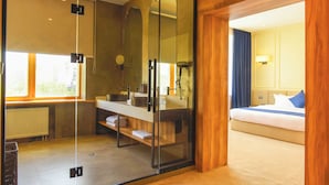 A modern hotel room with a glass-enclosed shower, a wooden floor, a bed with blue bedding, and a view of the city through large windows. - Ulaanbaatar Hotel (Ulaanbaatar)