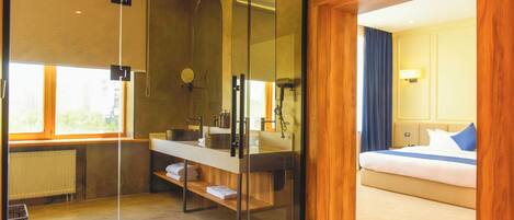 Una habitación de hotel moderna con ducha envuelta en vidrio, suelo de madera, cama con ropa de cama azul y vistas a la ciudad a través de amplias ventanas.