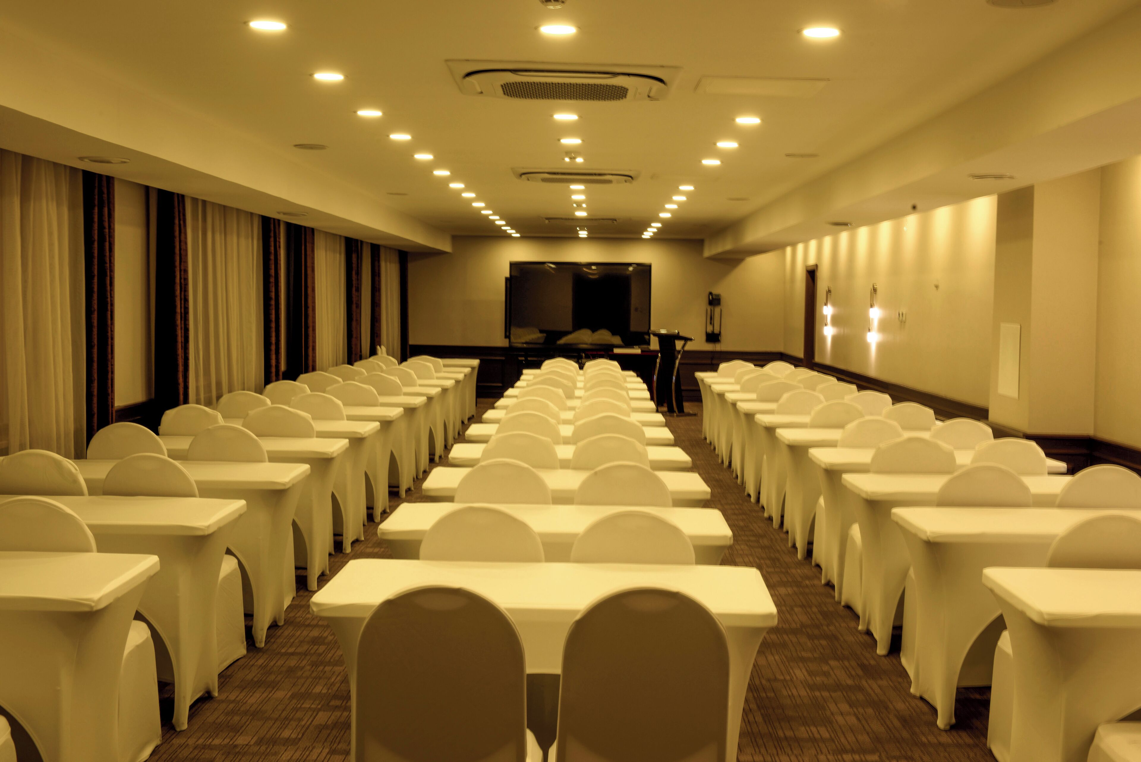 Une salle de conférence avec des rangées de tables et de chaises blanches, un grand écran à l’avant et un sol recouvert de moquette.