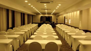 Un salón de conferencias con filas de mesas y sillas blancas, una pantalla grande al frente y piso alfombrado.