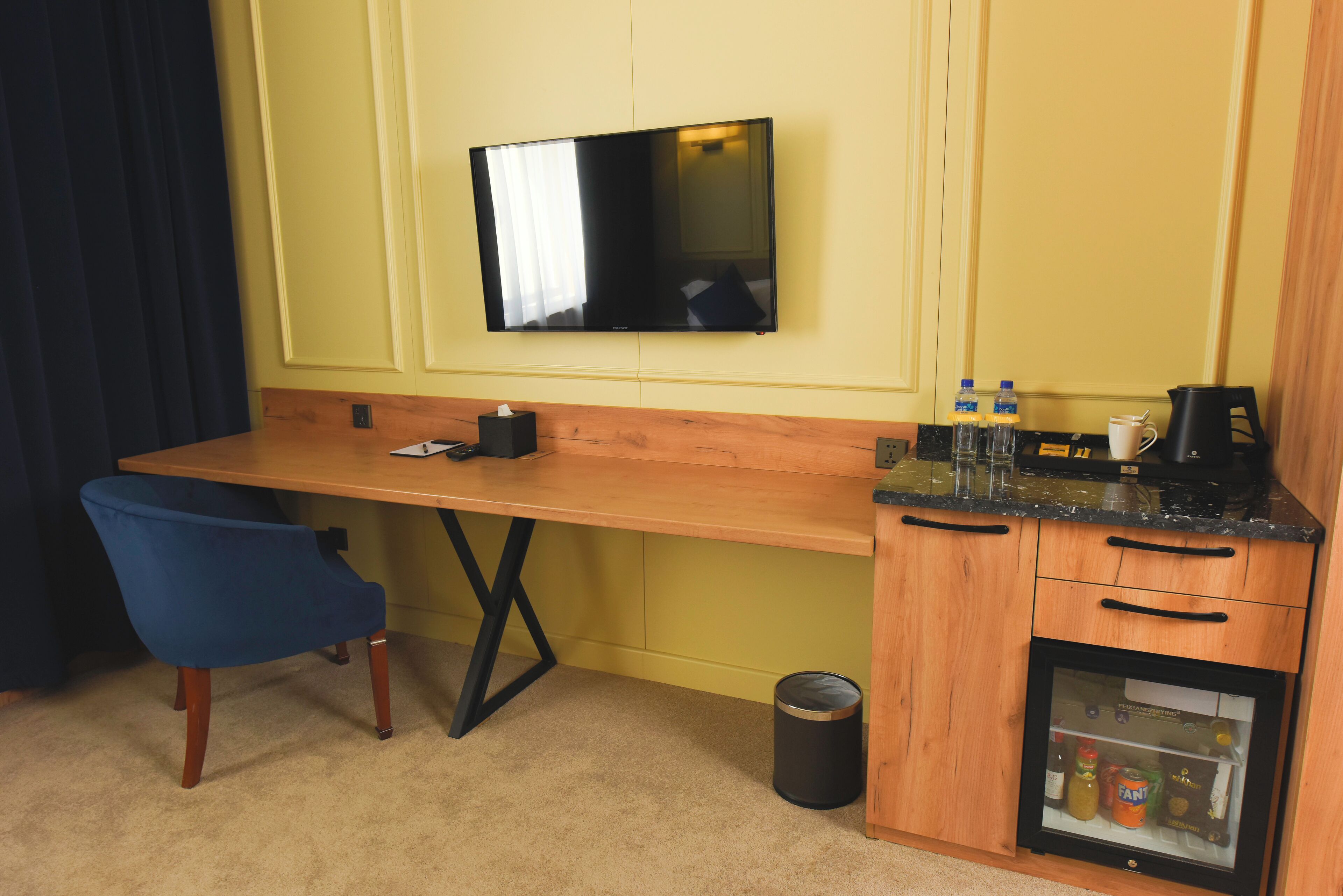 A compact hotel room with a wooden desk, a blue chair, a television mounted on the wall, a small refrigerator, and a coffee maker.
