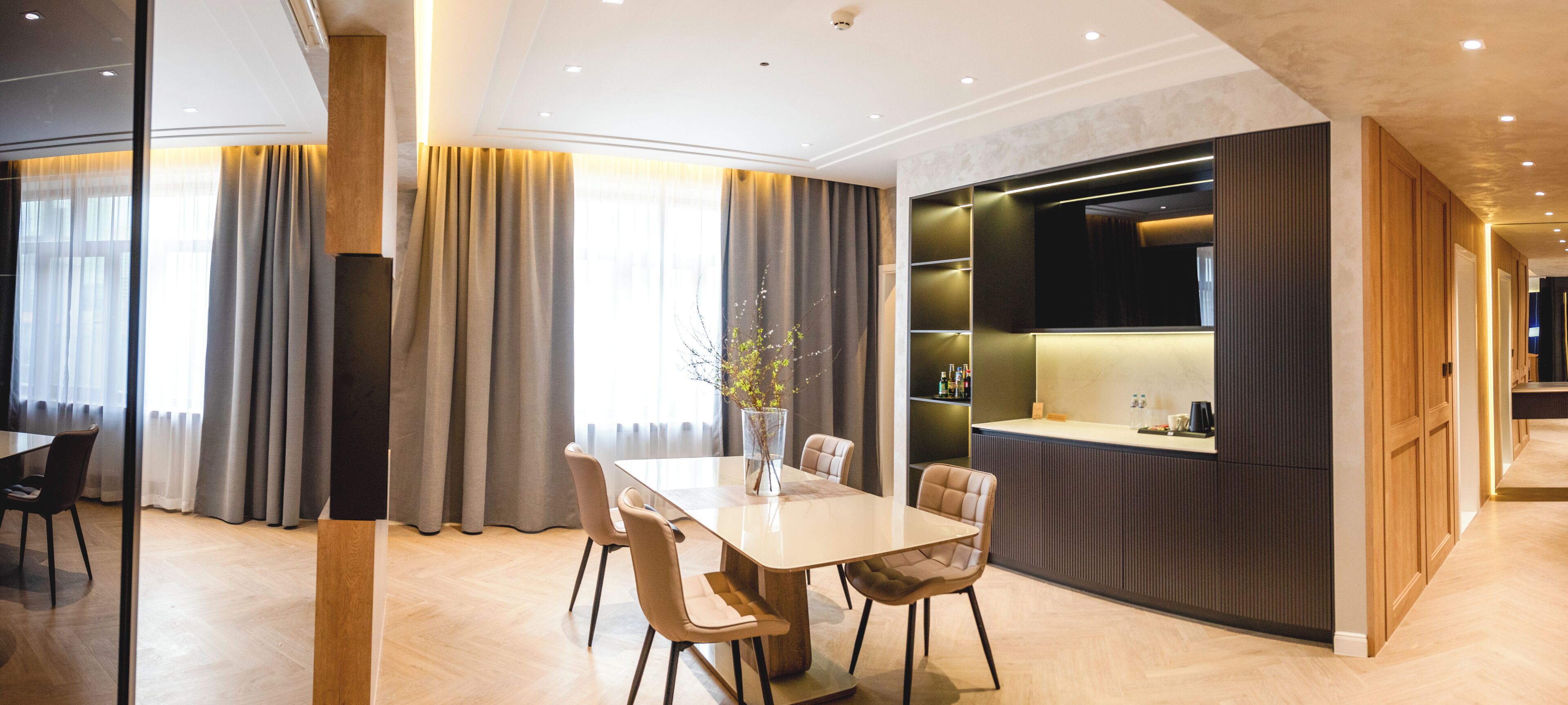 A modern hotel room with a dining area, a kitchen counter, and a seating area with a vase of flowers.