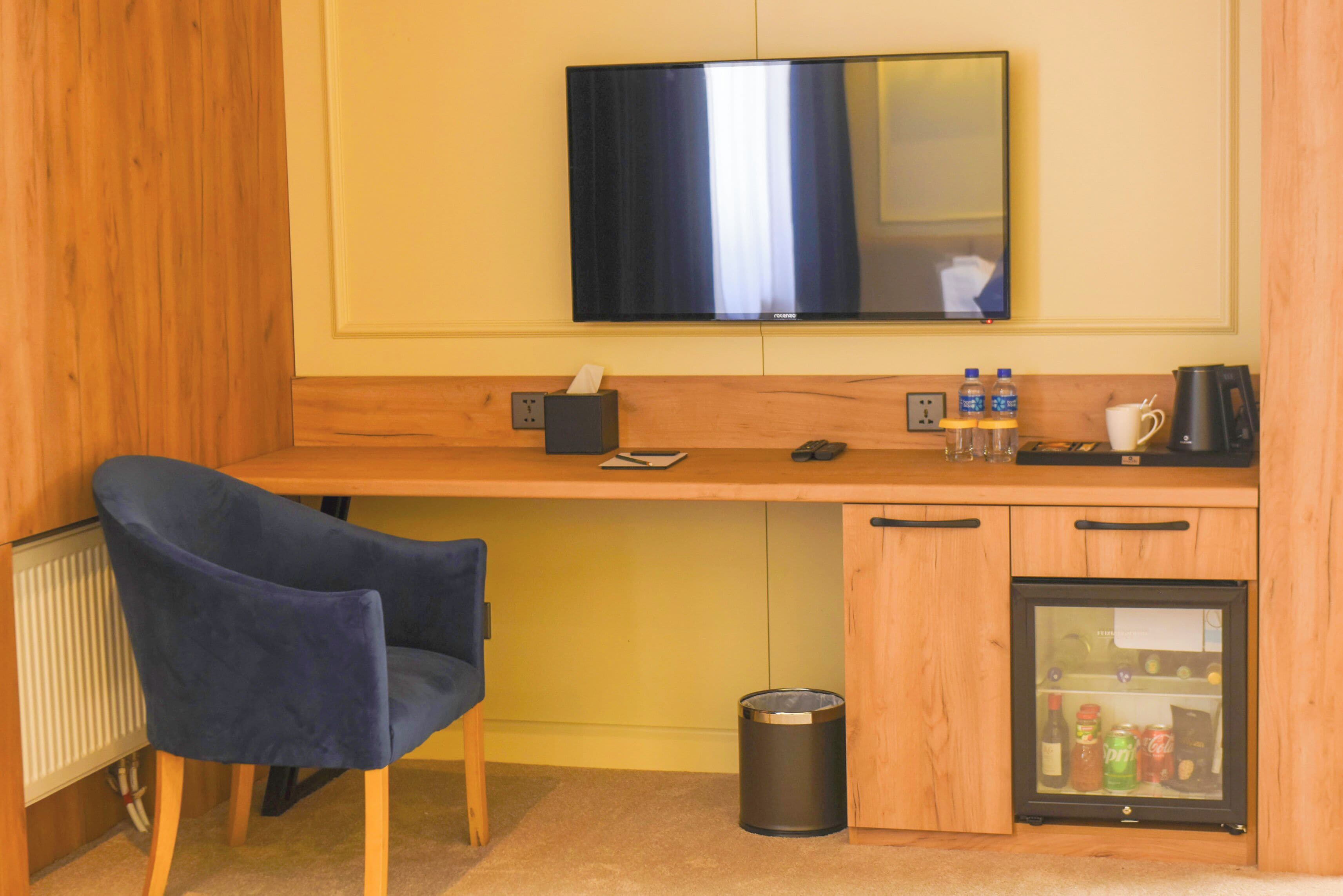 Habitación de hotel con un escritorio de madera, una silla azul, un televisor fijado a la pared, un refrigerador y un bote de basura.