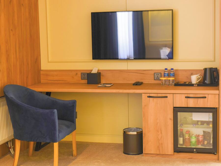 A hotel room with a wooden desk, a blue chair, a television mounted on the wall, a refrigerator, and a trash can.