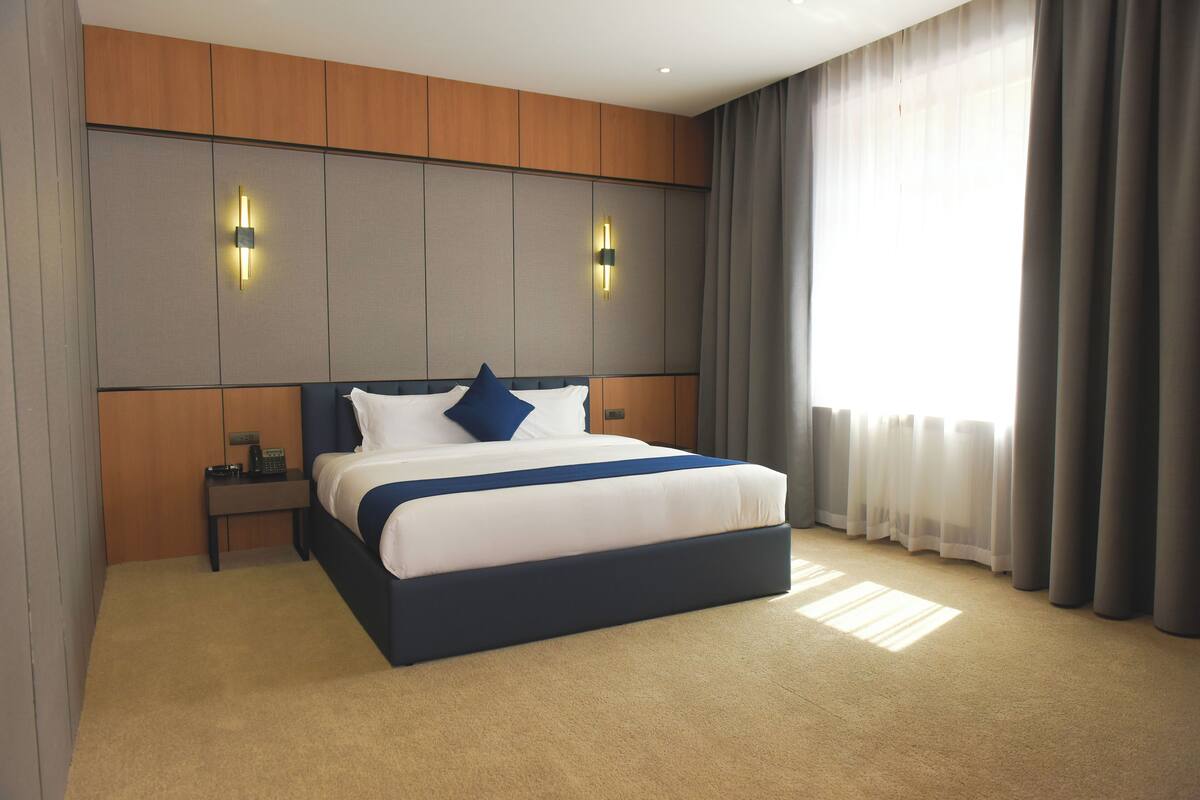 A modern hotel room with a large bed, two bedside tables, a headboard with built-in lighting, and a large window with curtains.