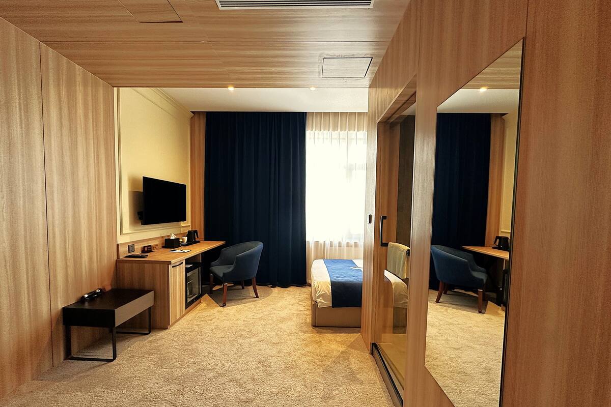 A hotel room with a wooden ceiling, a flat-screen TV, a desk with a chair, a bed with a blue blanket, and a large mirror.