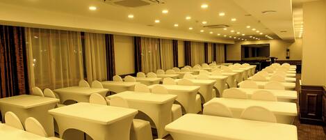 A conference room with rows of white tables and chairs, a large flat-screen TV, and a ceiling with recessed lighting.