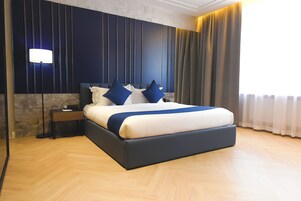 Quarto de hotel moderno com uma cama grande, painéis de parede azul escuro, uma luminária de cabeceira e vista para o exterior.