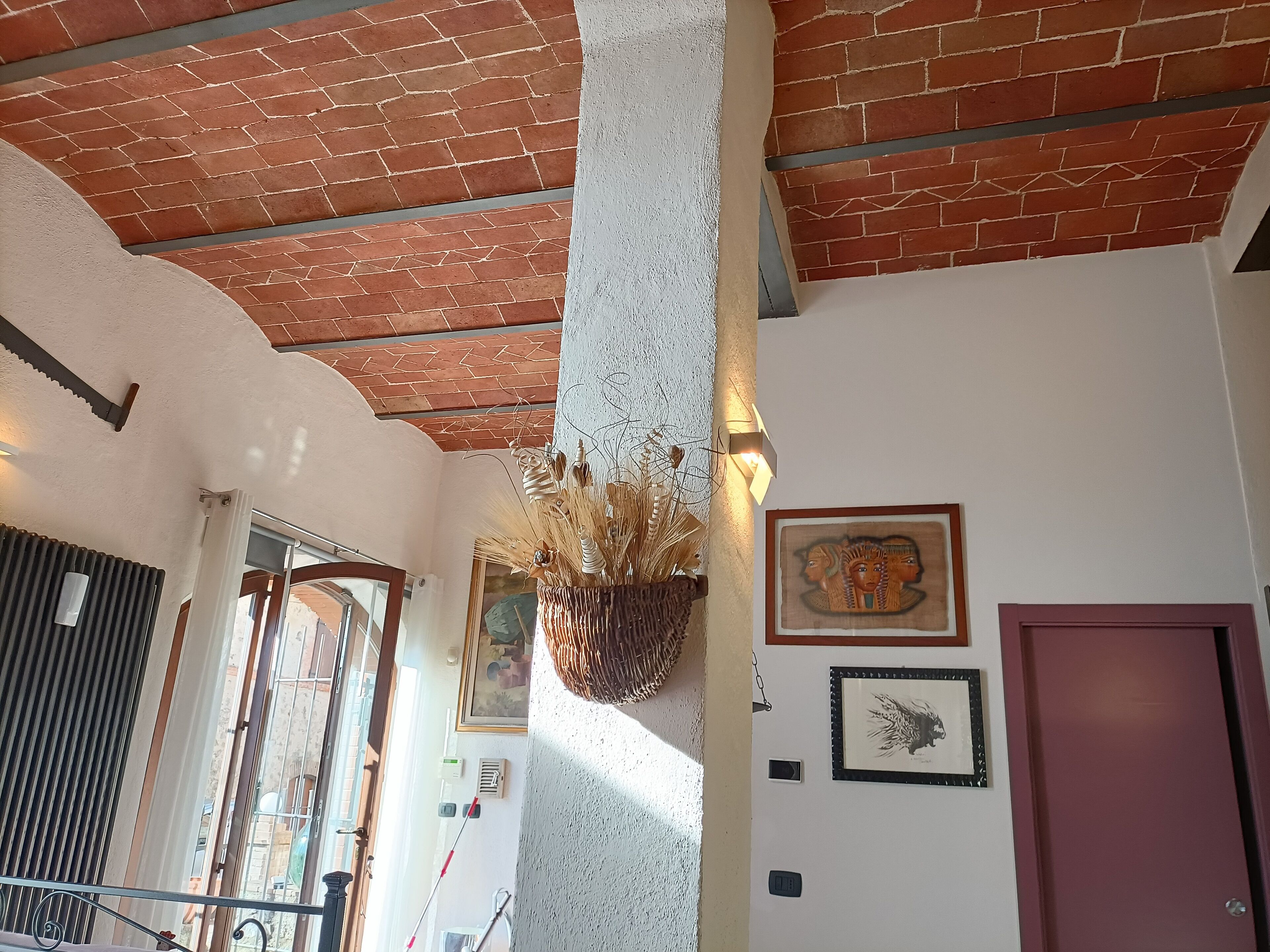 Een kamer met een bakstenen plafond, een witte kolom, een hangende plant en lijstwerken aan de muren.