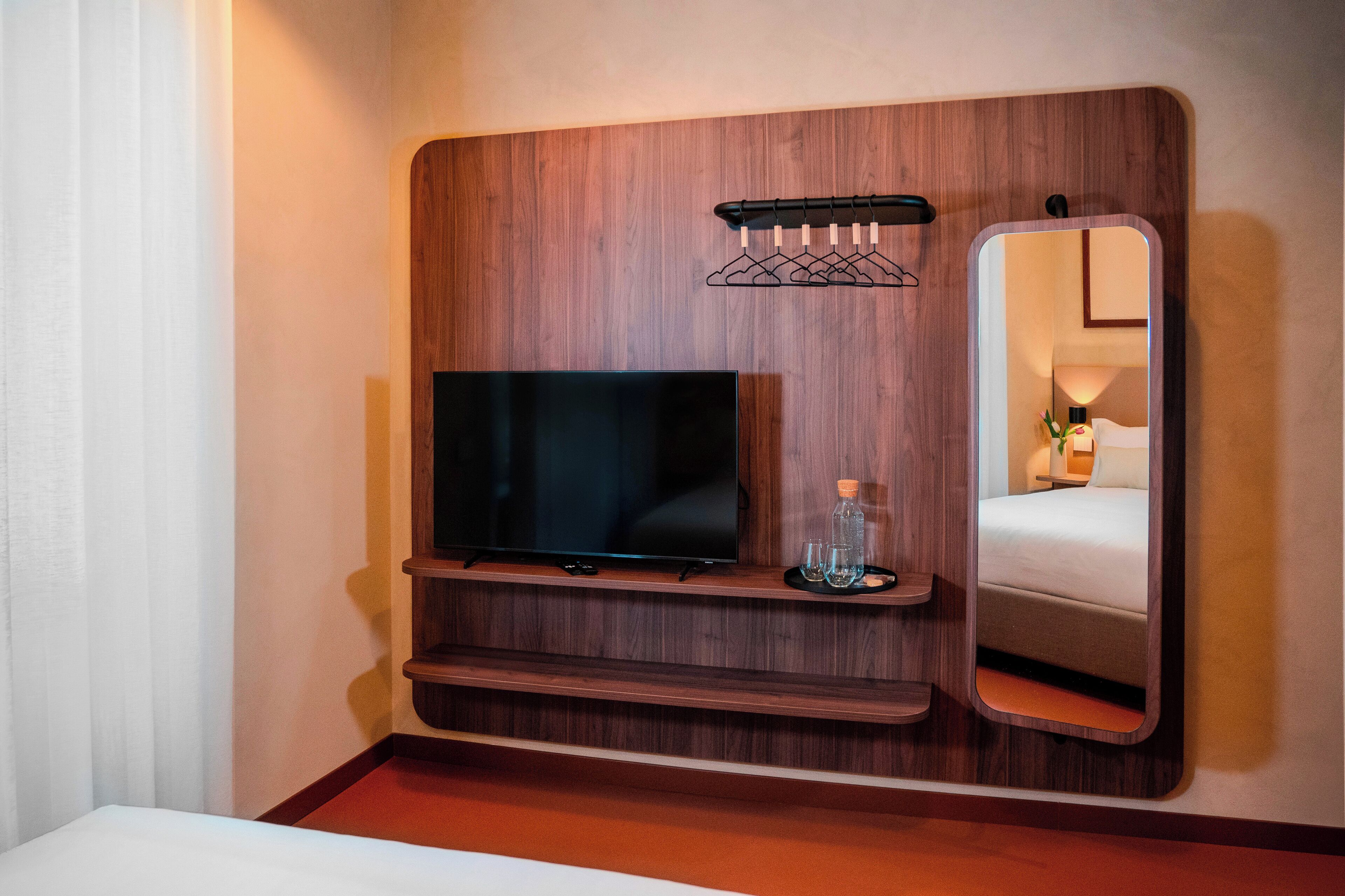 Habitación de hotel con pared de paneles de madera, televisor de pantalla plana, una pequeña estantería con vasos y una botella, y un espejo.