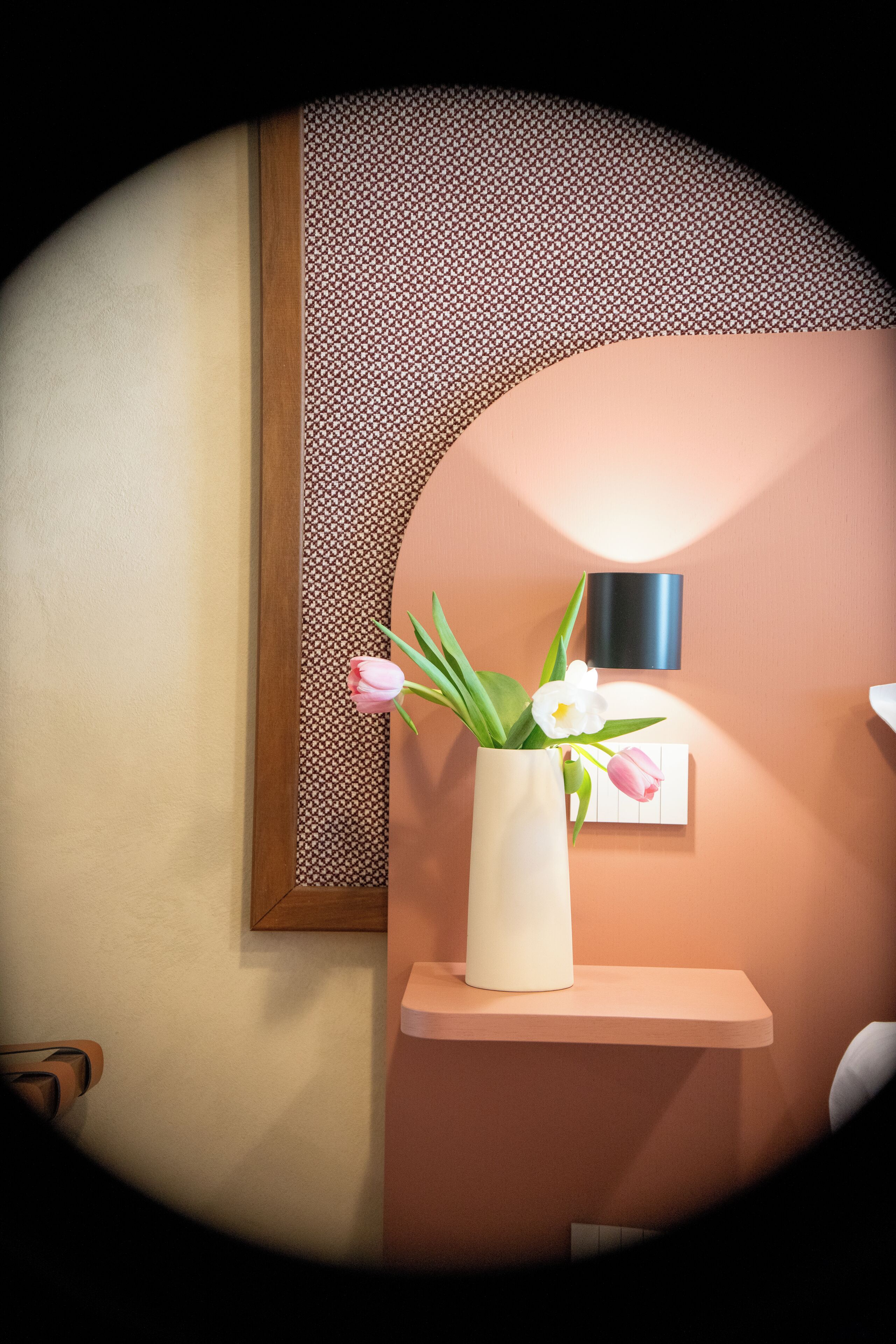 A bathroom with a white vase containing flowers on a shelf, a wall-mounted light, and a framed mirror.