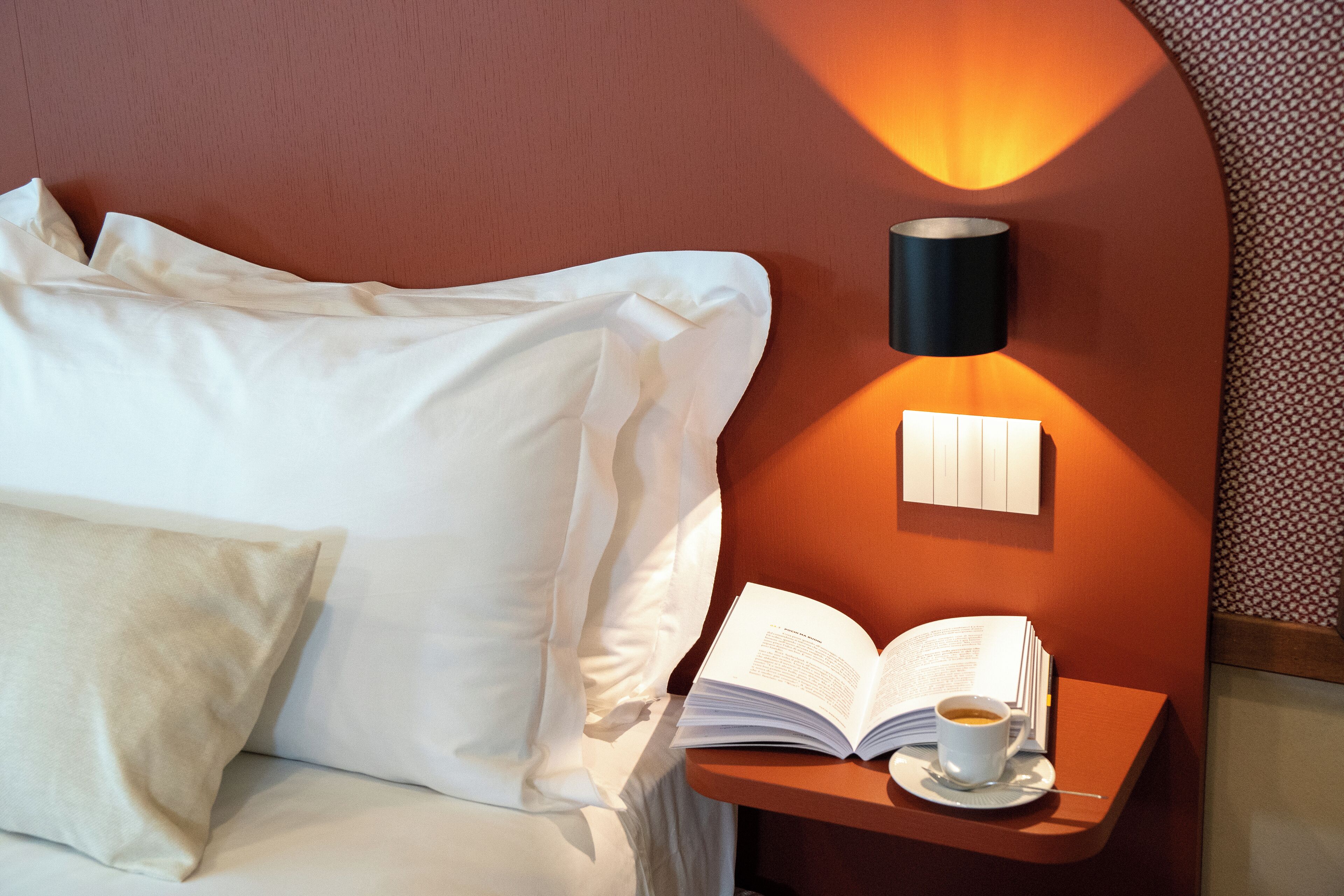A neatly made bed with white linens and pillows, a bedside table with a book and a cup of coffee, and a wall-mounted lamp.