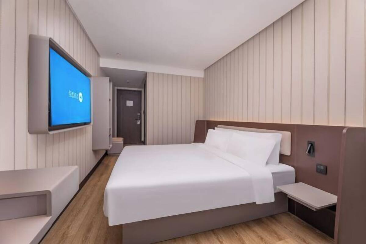 A modern hotel room with a large bed, a TV mounted on the wall, a bedside table, and a wooden floor.