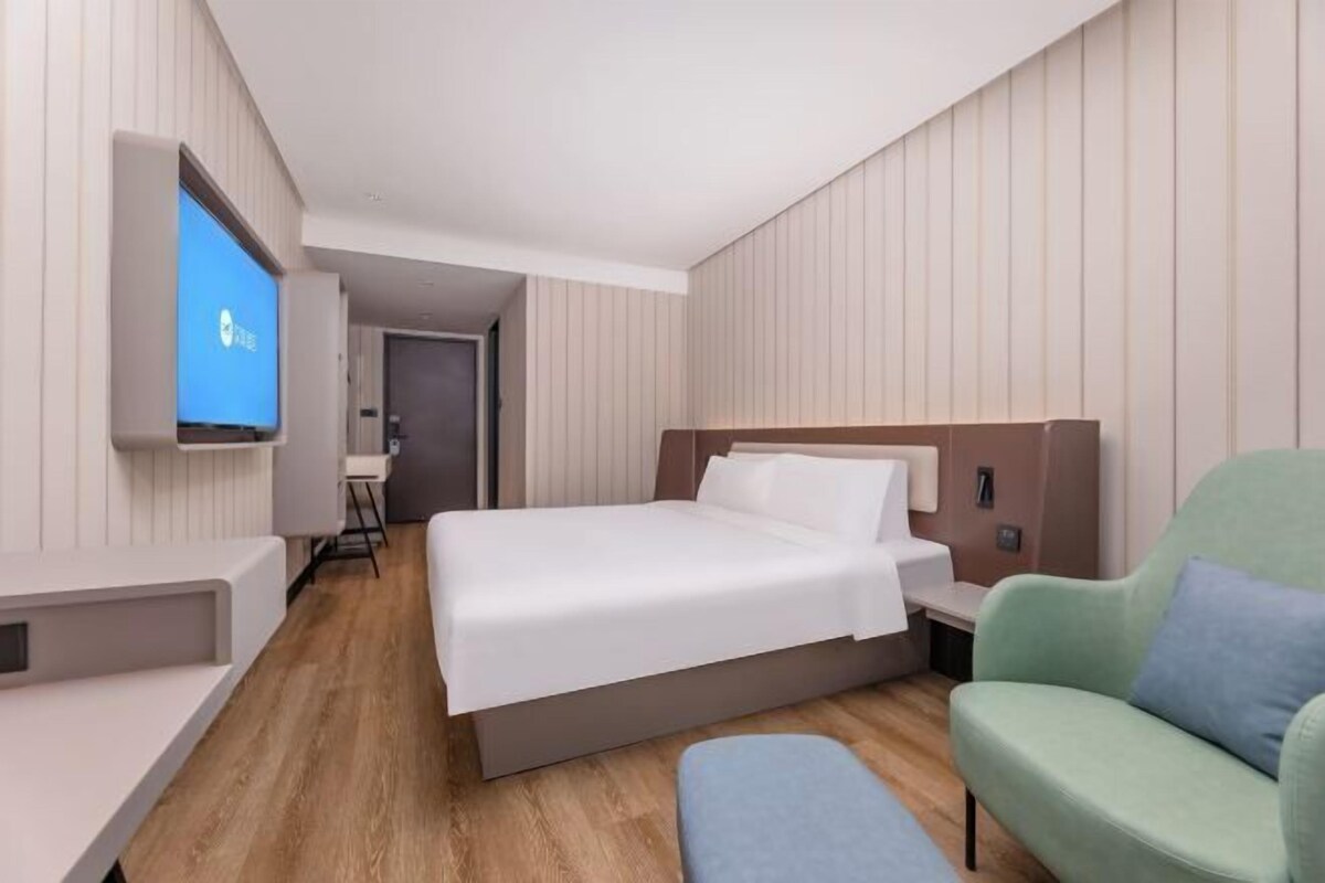 A modern hotel room with a large bed, a TV mounted on the wall, a desk, and a green armchair.
