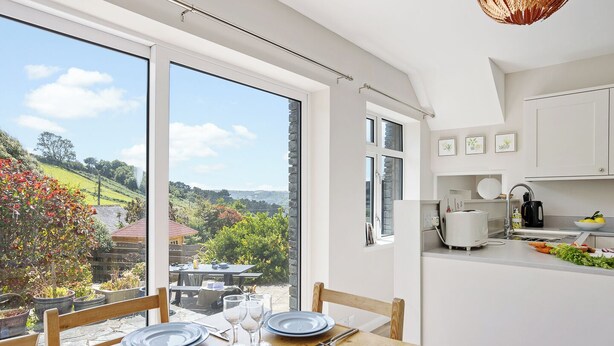 Une cuisine moderne avec des meubles blancs, un coin repas doté d’une table en bois et une grande fenêtre offrant une vue imprenable sur un paysage verdoyant.