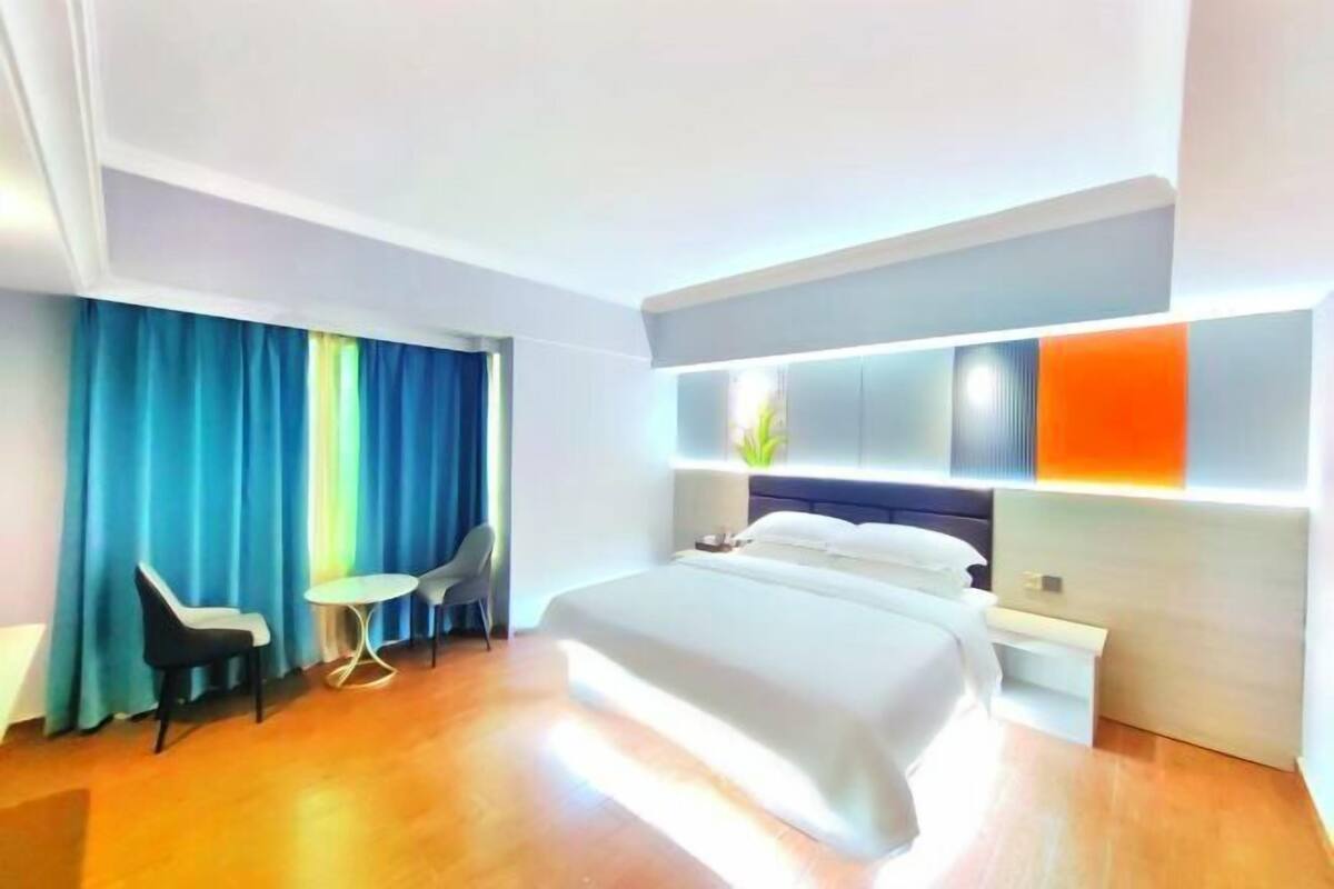 A modern hotel room with a bed, bedside tables, a small round table, two chairs, and a window with curtains.