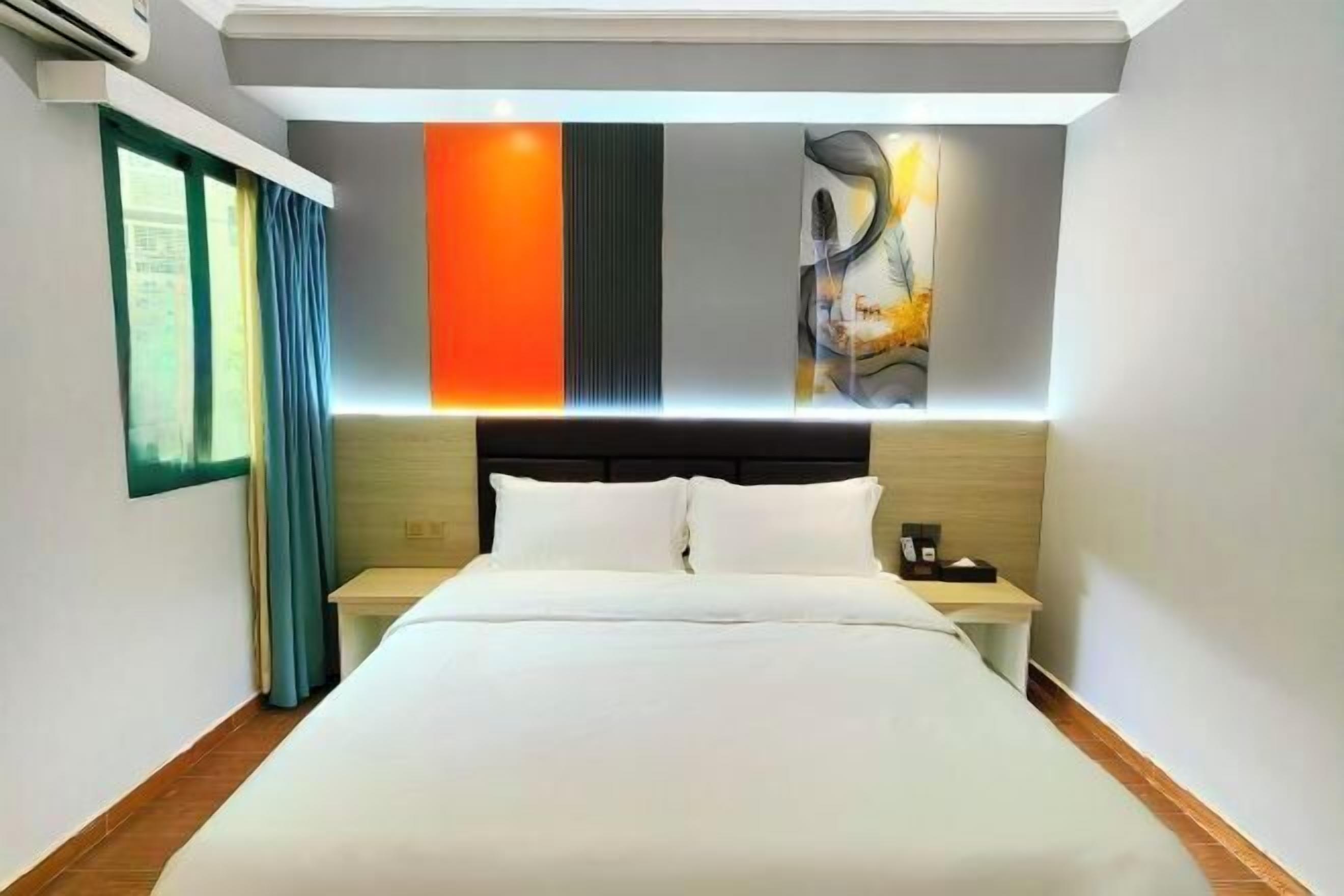 A hotel room with a large bed, a nightstand, a wall-mounted air conditioner, and a window with curtains.