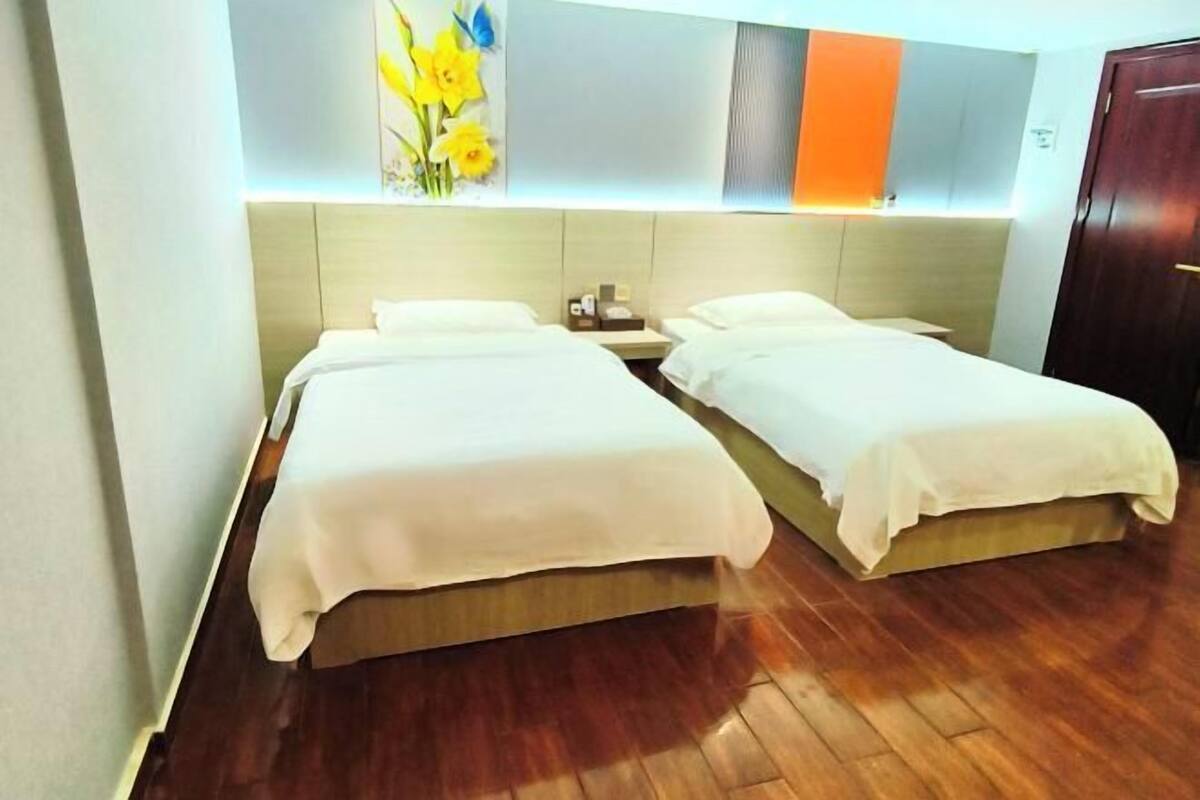 A hotel room with two single beds, a wooden floor, a wall-mounted light fixture, and a door.