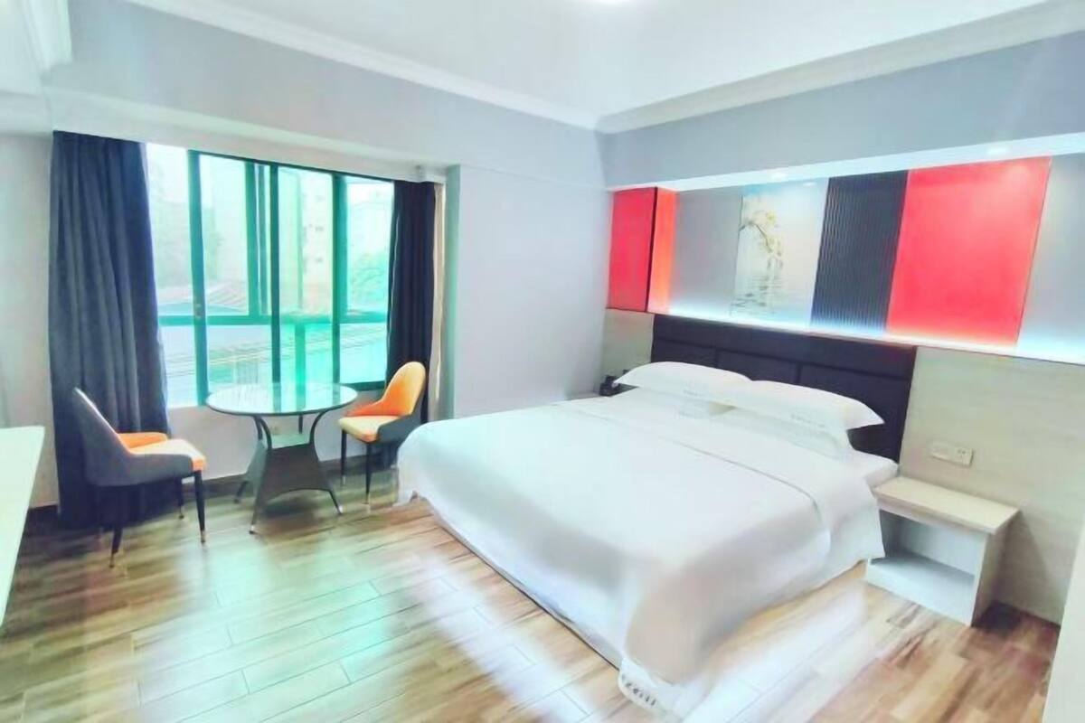 A hotel room with a large bed, a small table, two chairs, a window with curtains, and a modern wall design.