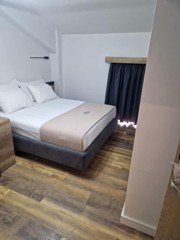 A bedroom with a bed, wooden flooring, a nightstand, and a window with curtains. - Lumi i Bardhe Hotel & Restaurant (Horë-Vranisht)