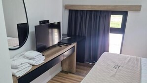 A hotel room with a bed, a desk with a computer, a mirror, and a window with curtains.
