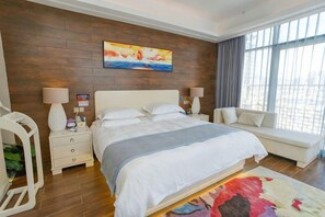 Une chambre d’hôtel moderne, dotée d’un grand lit, de tables de chevet, d’un canapé et offrant une vue sur l’extérieur grâce à de grandes fenêtres.