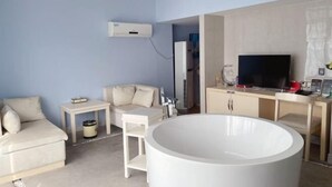 Moderni hotellihuone, jossa on beigenvärinen sohva, pieni valkoinen pöytä, valkoinen kylpyamme, televisio ja minibaari.