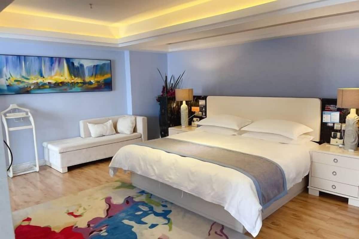 A bedroom with a large bed, a bedside table, a nightstand, a bench, a painting, and a floor lamp.