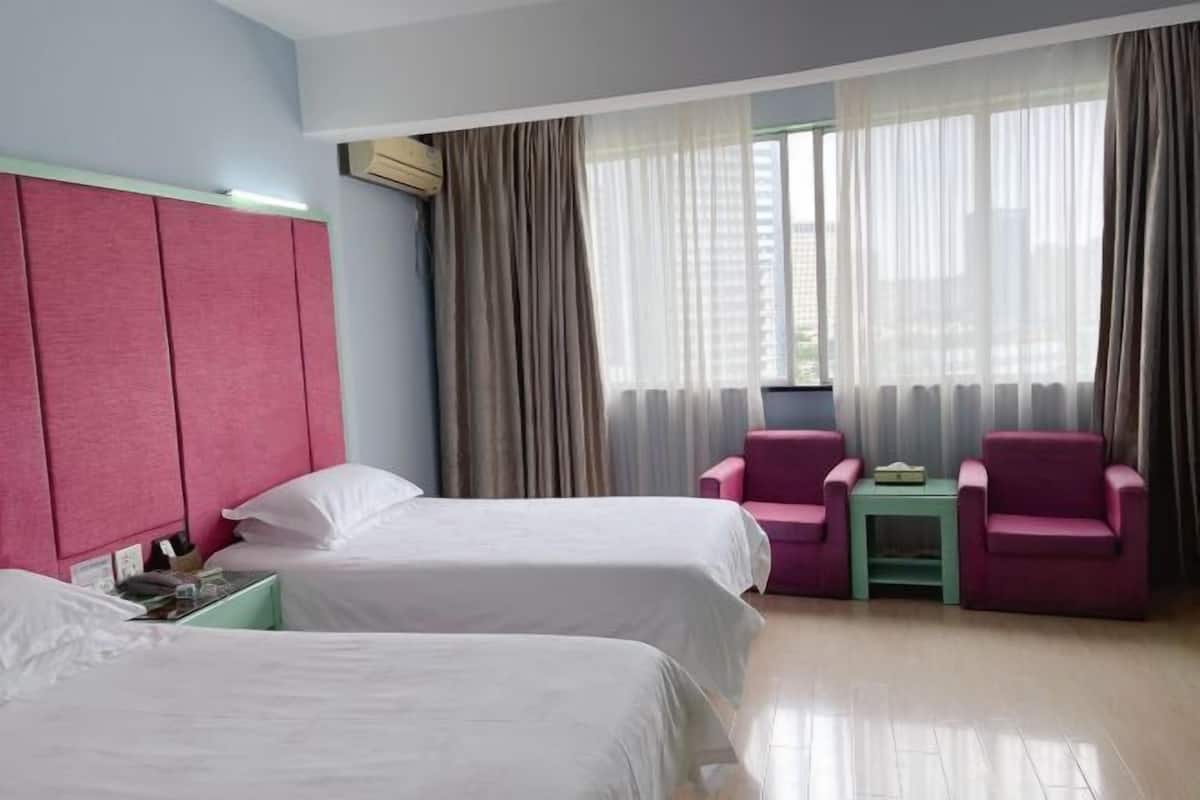 A hotel room with two beds, a pink headboard, a small table with chairs, and a window with curtains.