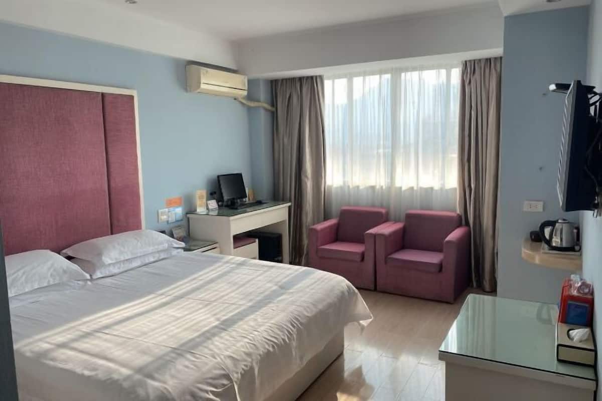 A hotel room with a bed, a desk with a computer, two chairs, a TV, and a window with curtains.