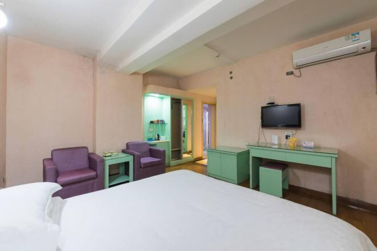 A hotel room with a bed, two chairs, a desk, a television, and an air conditioning unit.