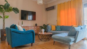 Ein Wohnzimmer mit blauem Sofa, grauem Sofa, Couchtisch aus Holz, einer großen Pflanze und einem flachbildfernseher an der Wand.