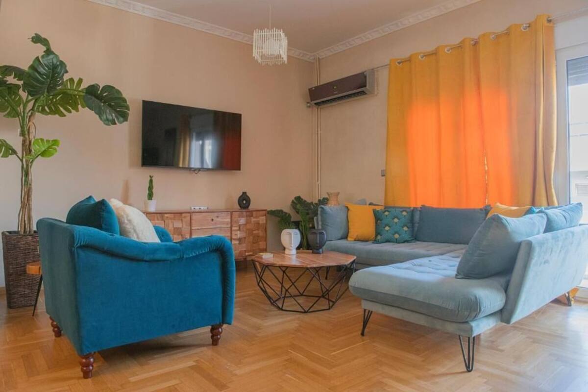 A living room with a blue sofa, a grey sofa, a wooden coffee table, a large plant, and a flat-screen TV mounted on the wall.