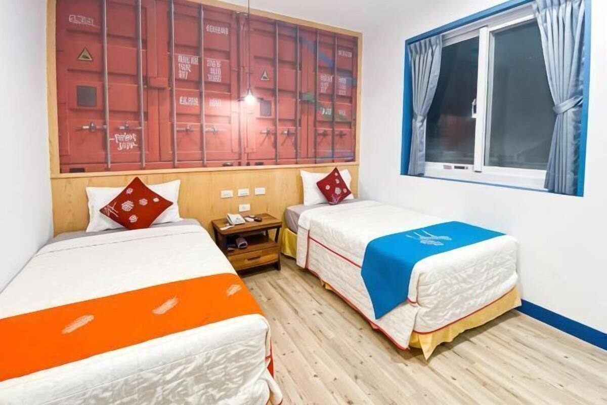 A hotel room with two beds, a wooden floor, a window with curtains, and a wall with storage lockers.