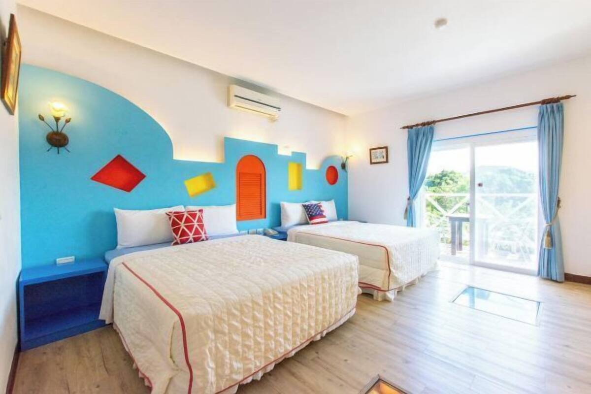 A room with two beds, a blue accent wall, and a large window with curtains.