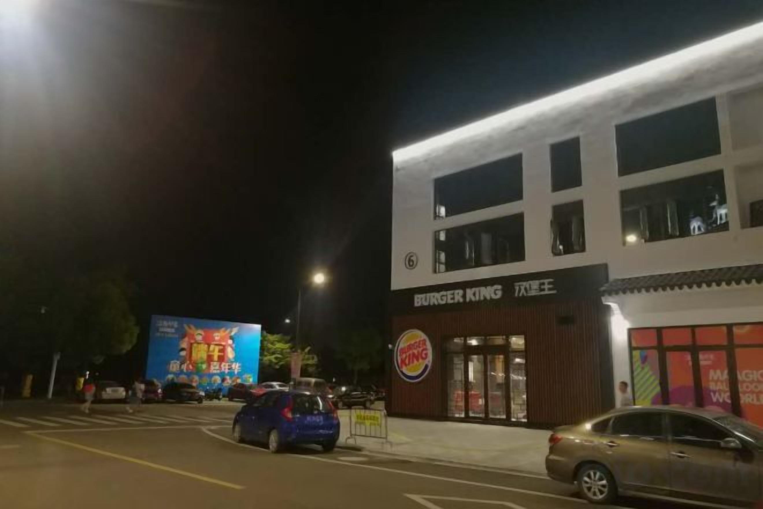 Ein Burger King Restaurant bei Nacht mit einer großen, beleuchteten Schautafel, parkenden Autos und einer Werbetafel im Hintergrund.