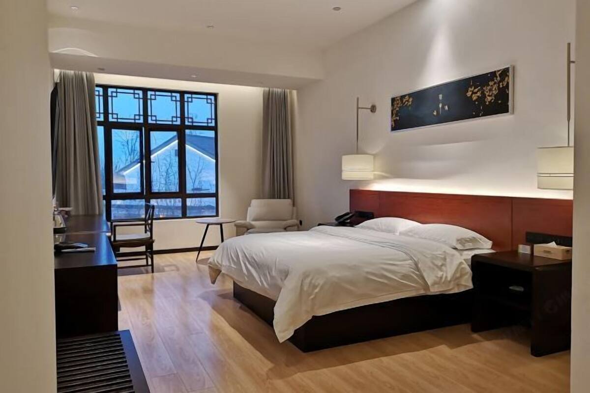 A modern hotel room with a large bed, a desk, a chair, a bedside table, a lamp, and a window with curtains.